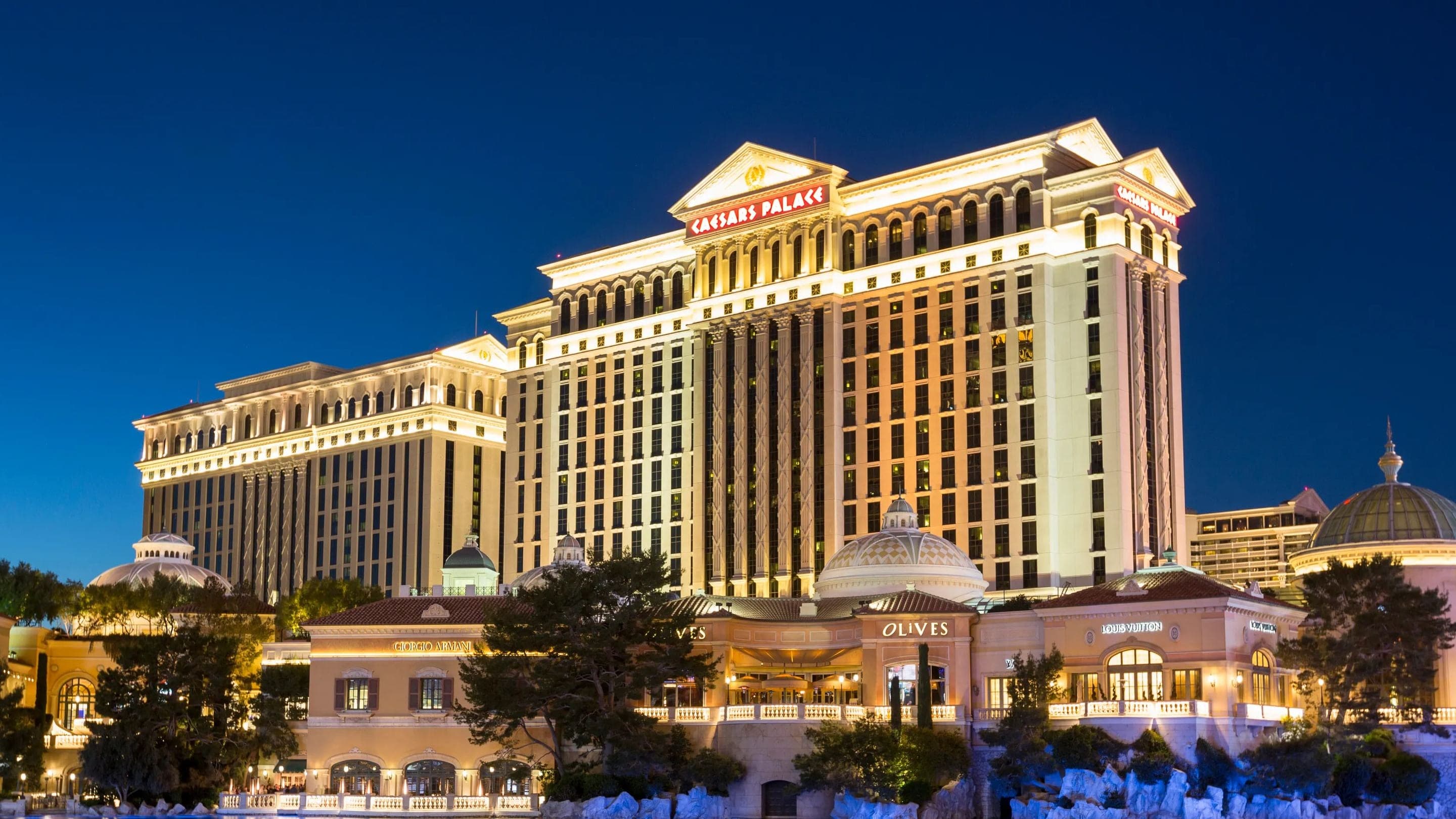 Caesars Palace hotel towers lit up at night on the Las Vegas Strip, reflected in the Bellagio fountains lake